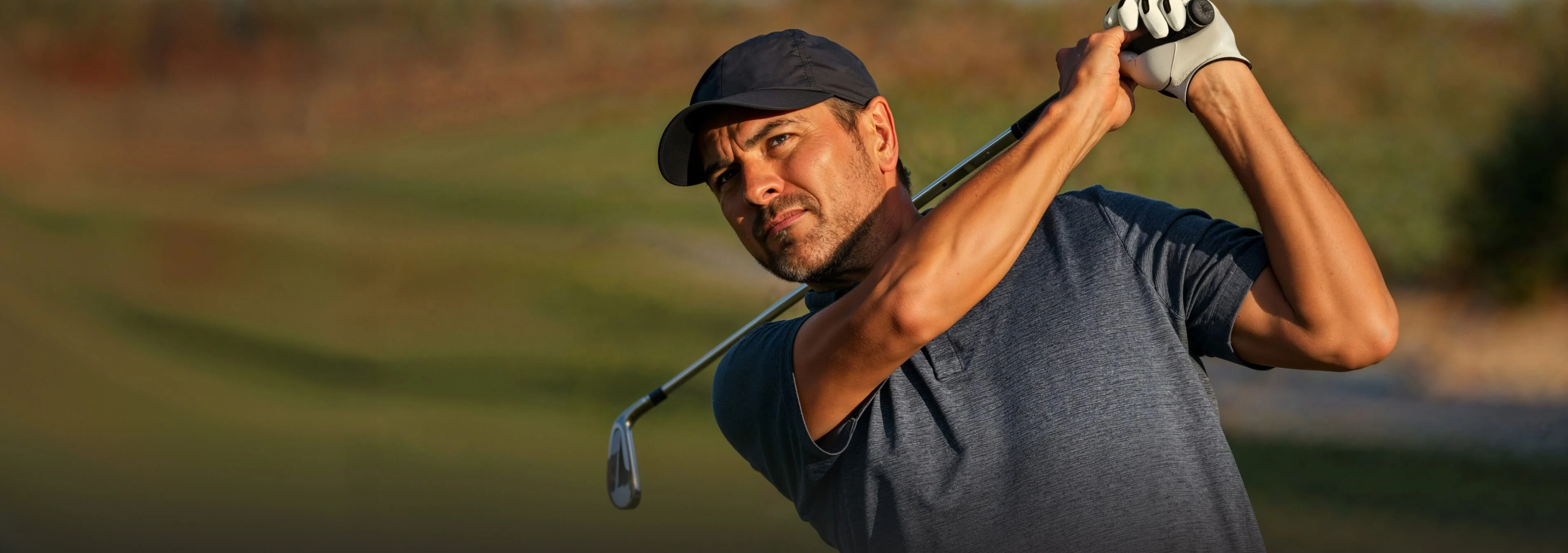 A man wearing a black T-shirt and black cap holds a golf club, preparing to take a swing on a green field.